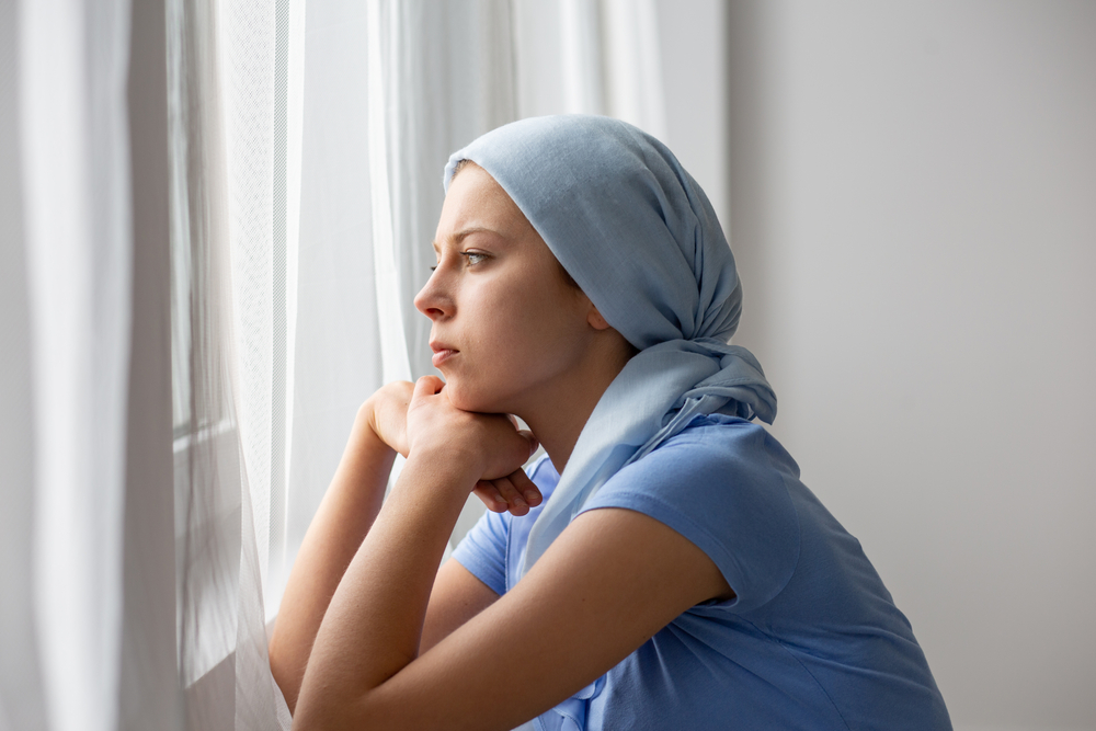 Thoughtful young girl suffering from ovarian cancer, wearing blue headscarf and looking through the window in medical center